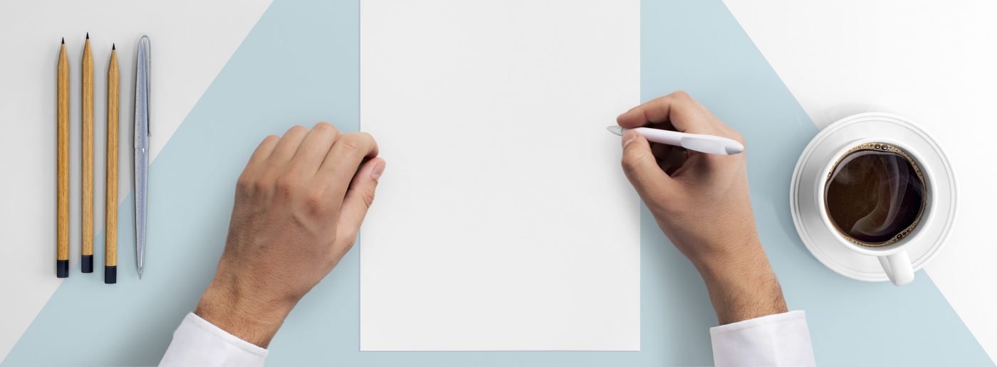 Direct view at hands writing on a blank paper .Office desk with pc keyboard, smartphone, coffee, notebook, pencils, white and blue desk pattern. copy space. hipster template