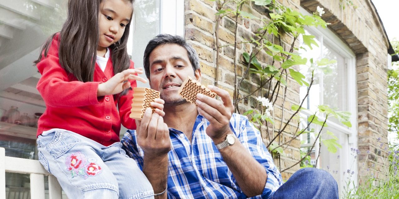 Man and girl inspecting miniature clay blocks in front of brick-lined house