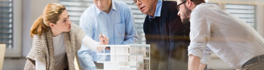 Architect explaining project plan to client in office with the help of an architectural model. Project manager and co-worker standing beside him.