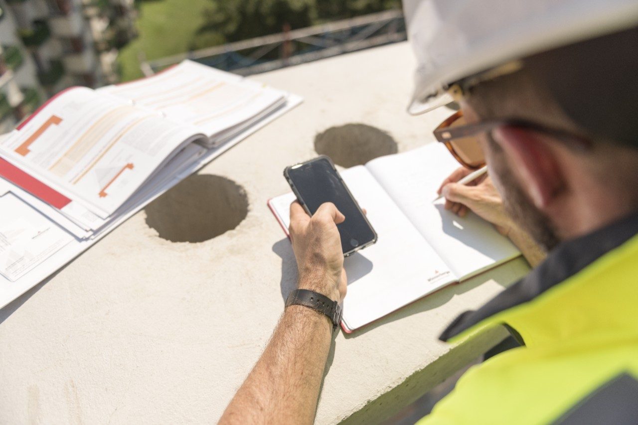 Urban roofer taking notes holding iPhone and writing in an urban location wearing hard hat and safety jacket