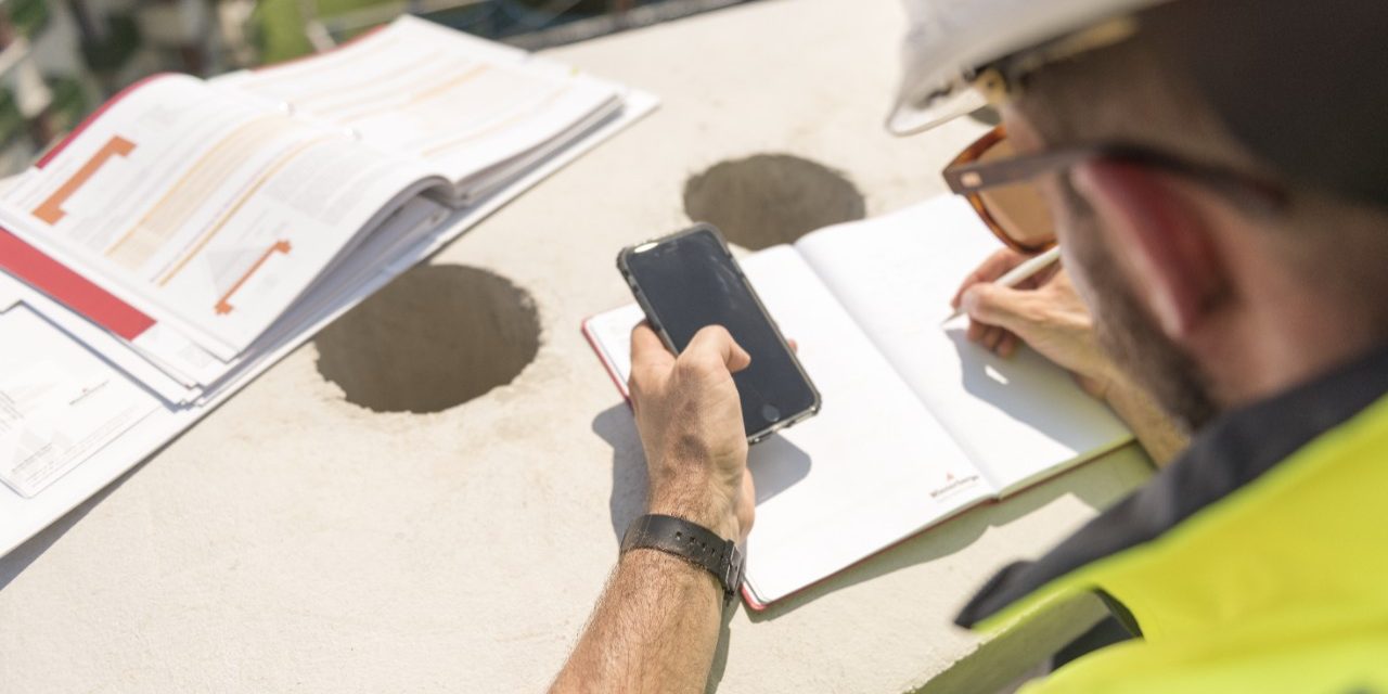 Urban roofer taking notes holding iPhone and writing in an urban location wearing hard hat and safety jacket