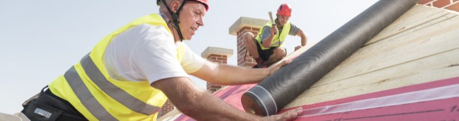 Urban roofers applying roof underlay sheet wearing hard hat and safety jacket brick chimney