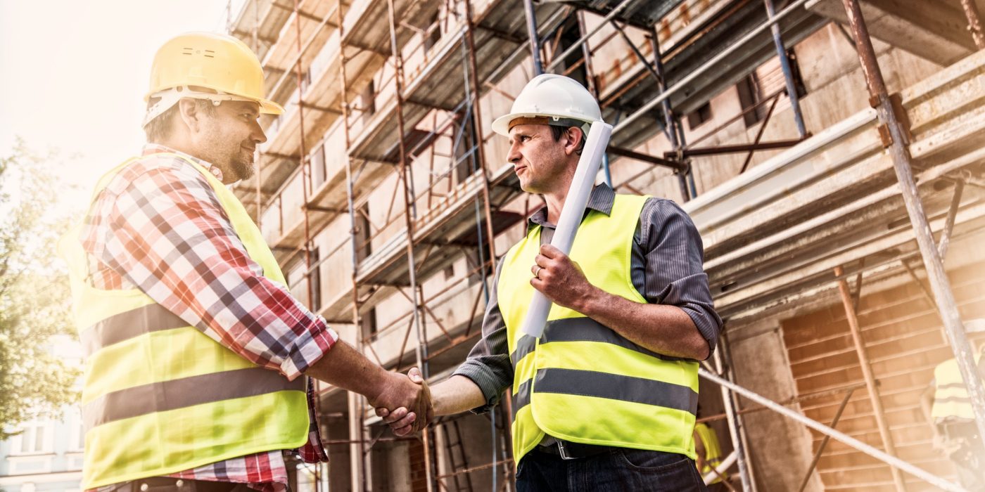 Handshake of two man wearing reflective clothing and work helmets o a construction site, scaffolding in background.