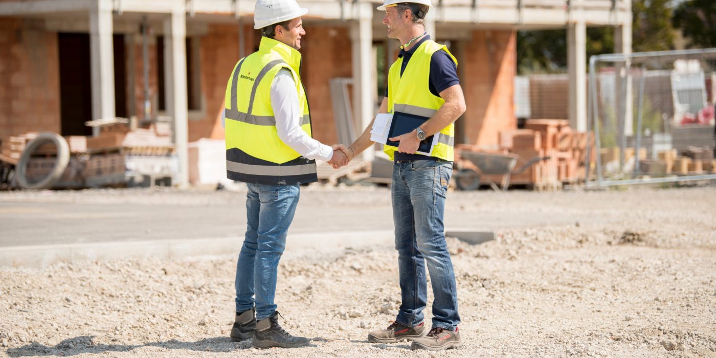 Manager and builder shaking hands on construction site in front of building shell