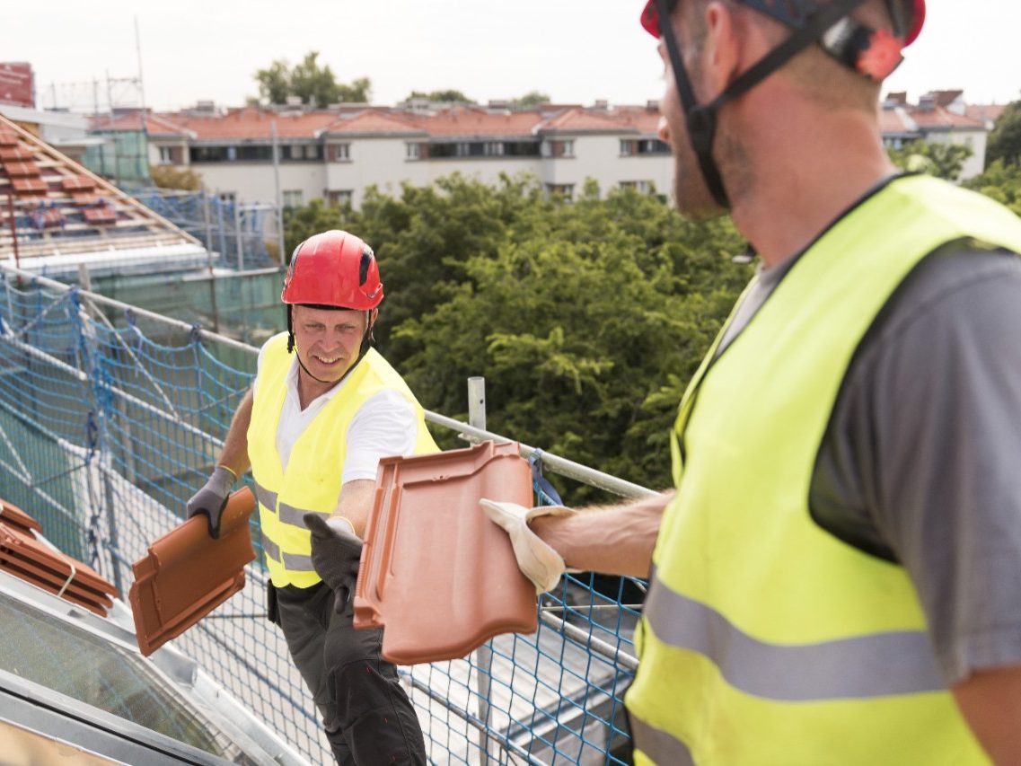 Urban roofers passing roof tile towards colleague roofing with safety jackets and hard hats