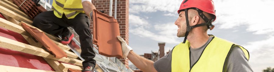Urban roofers passing on roof tile towards colleague working on the roof wearing safety jackets and hard hats